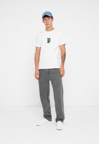 Hvit bomulls t-skjorte med en svart og neongrønn grafisk logo, paret med grå denimbukser og hvite sneakers, brukt med en lyseblå caps.