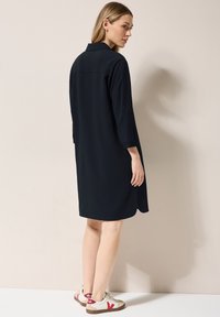 Abito camicia blu navy con colletto, maniche lunghe arrotolate e una texture liscia. Abbinato a sneakers beige e rosse.