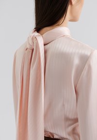 Light pink, satin blouse with vertical stripes. Features a tied scarf at the neckline and smooth texture with subtle sheen.