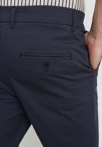 Granatowe spodnie wykonane z miękkiego materiału, z tylną kieszenią zapinaną na guzik oraz gładkim, czystym wzorem.