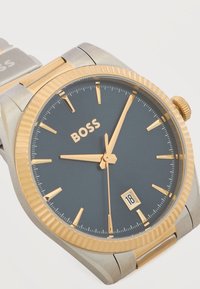 Ανδρικό ρολόι χειρός με χρυσό περίβλημα και δείκτες, μπλε καντράν που δείχνει ώρα 9:03:06, παράθυρο ημερομηνίας στη θέση 4, και δίχρωμο μεταλλικό λουράκι.