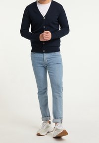 Mörkblå stickad kavaj med V-ringning, knappstängning och ribbade ärmavslut, tillsammans med ljusblå uppvikta jeans och vita sneakers med bruna detaljer.