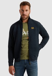 Marineblauer, gesteppten Jacke mit hohem Kragen, Reißverschluss vorne und elastischen Bündchen. Verfügt über ein strukturiertes Muster und ein gelbes Markenetikett auf der Brust.