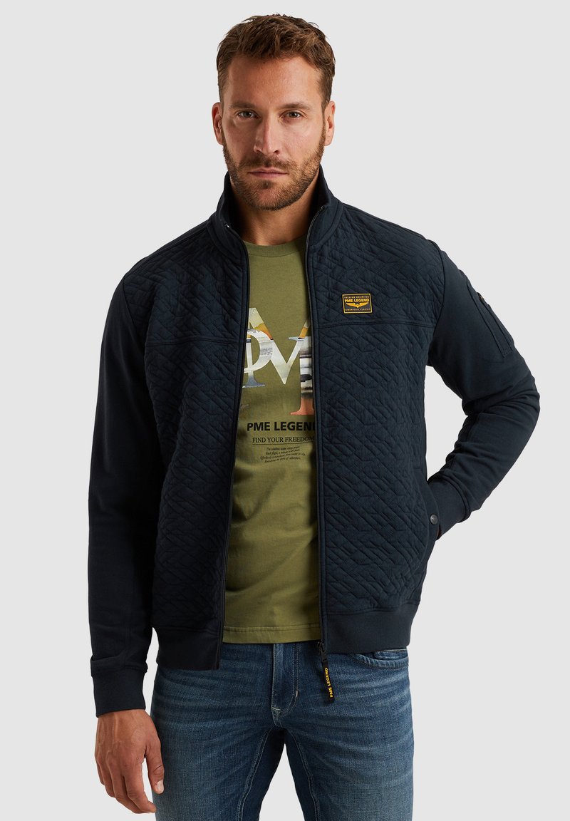 Marineblauer, gesteppten Jacke mit hohem Kragen, Reißverschluss vorne und elastischen Bündchen. Verfügt über ein strukturiertes Muster und ein gelbes Markenetikett auf der Brust.