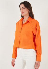 Bright orange cropped button-up shirt featuring a pointed collar, a single patch pocket, and long sleeves, made from a lightweight fabric.