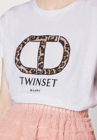 Bílý tričko s leopardím potiskem loga "TD" a textem "Twinset Milano", v kombinaci s růžovou sukní s volány.