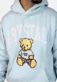Felpa blu chiaro con grafica di un orsetto seduto che indossa una maglietta "Crystal Paris", con testo bianco audace sopra.