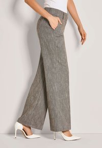 Vrouw in hoge, wijde grijze broek met een gestructureerd patroon, gecombineerd met witte puntige hoge hakken, poserende opzij.