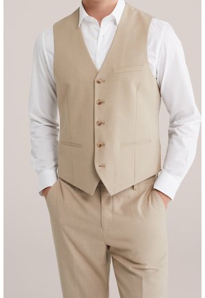 Man draagt beige knoopvest en bijpassende broek met wit overhemd met lange mouwen en kraag, handen in de zakken, neutrale achtergrond.