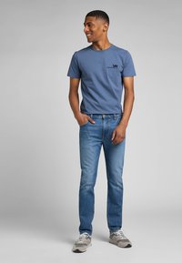 Męska niebieska koszulka z krótkim rękawem i okrągłym dekoltem, z czarnym logo, w połączeniu z jasnoniebieskimi dżinsami i szarymi trampkami. Prosty design.