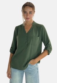 Blusa verde leggera con scollatura a V, apertura con bottoni e tasca sul petto. Presenta maniche arrotolate e una vestibilità comoda.