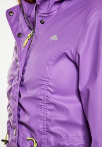 Purpurinė vandeniui atspari striukė su blizgiu paviršiumi, turinti raukšlėtą dizainą, sidabrinį skėčio logotipą ir geltonus akcentus užtrauktukuose bei stygose.