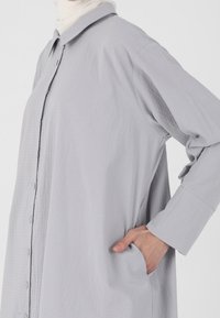 Chemise gris clair à manches longues avec col, à la texture originale et à la coupe décontractée. Comprend des poignets et des poches latérales.