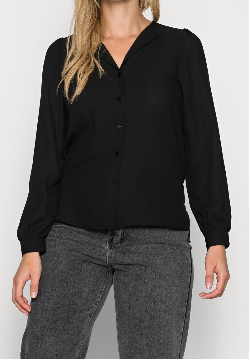 Femme portant un chemisier noir boutonné à manches longues et un jean gris, recadré au niveau du cou et de la moitié de la cuisse.