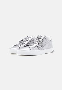 Srebrne metalik tenisice s okruglim vezicama, bijelim gumenim potplatom, reljefnim logotipom sa strane i okruglim metalnim detaljem. Glatka tekstura.