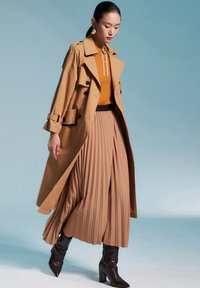 Manteau trench tan par-dessus un polo côtelé orange et une jupe plissée beige, associé à des bottes à talons hauts en cuir de crocodile noir.