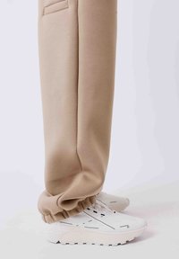 Beige joggingbroek met boord aan de enkel, gecombineerd met witte sportschoenen met zwarte lijnaccenten en een dikke zool.