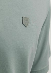 Zelená tričko s krátkým rukávem z měkké látky, které má texturované vyšívané logo štítu na levé straně hrudi.