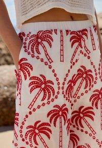Hvide bukser har rød palme-træs broderi med cirkulære detaljer. Livremmen er elastisk, og stoffet ser let og struktureret ud.