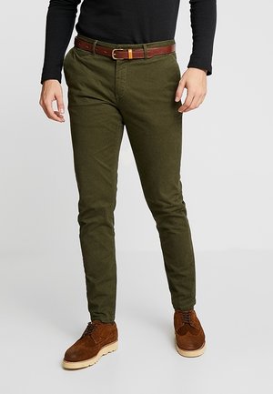 Mann trägt olivgrüne Slim-Fit-Hose mit braunem Ledergürtel, schwarzes Langarmhemd und braune Wildlederschuhe.