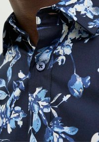 Camicia navy a fiori con motivi floreali blu e bianchi, colletto abbottonato e un unico bottone scuro decorativo sulla patta.