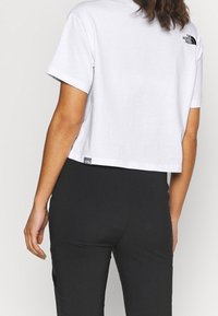Camiseta blanca corta de algodón con mangas cortas. Presenta un logo negro en la espalda y una etiqueta pequeña en el dobladillo. Combinada con pantalones negros.