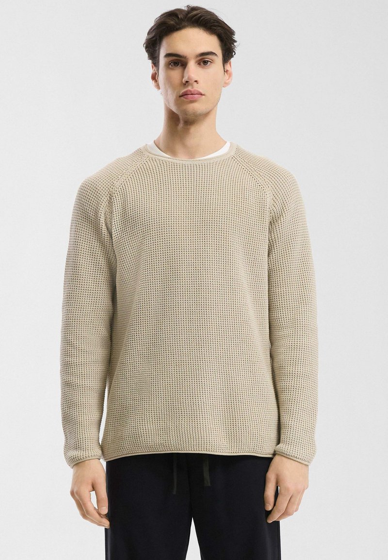 Beige gebreide trui met een textuur, geweven patroon, ronde halslijn en lange raglanmouwen, gedragen over een witte blouse, casual gestyled.