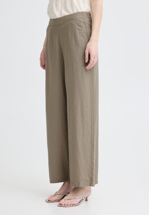 Personne portant un pantalon kaki ample à jambes larges avec des poches latérales et des sandales à talons beiges, debout devant un fond clair uni.