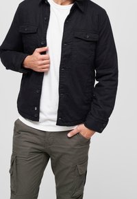 Svart knäppjacka med två bröstfickor och en lätt texturerad tyg, bärs över en vit T-shirt och olivgröna cargobyxor.