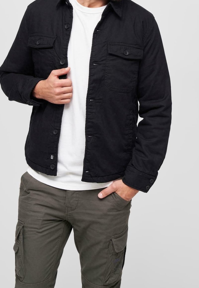 Svart knäppjacka med två bröstfickor och en lätt texturerad tyg, bärs över en vit T-shirt och olivgröna cargobyxor.