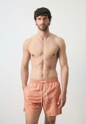 Hombre con cabello rizado oscuro y barba, vistiendo pantalones cortos de baño con patrón coral, de pie contra un fondo claro y liso.