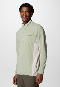 Uomo che sorride, indossando un pullover in fleece Columbia verde chiaro con zip a un quarto e pannelli laterali beige, in piedi contro uno sfondo grigio.