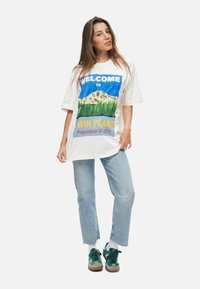 Witte oversized t-shirt met een grafisch ontwerp van bergen en bomen, met "WELKOM BIJ WIN PEAKS" in felle kleuren. Gecombineerd met lichte jeans en groene sneakers.