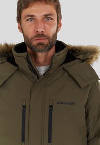 Parka imbottita verde oliva con cappuccio foderato in pelliccia, due tasche anteriori con zip e dettagli in metallo nero. Presenta una toppa sulla spalla per le regolazioni.