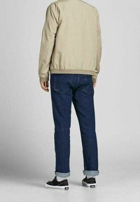 Béžová bomber bunda, modré džíny s ohrnutými manžetami a černé tenisky s bílými podrážkami, pohled zezadu. Hladká látka a volný střih.