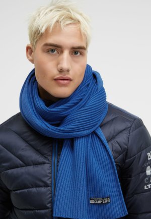 Blauer gestrickter Schal mit gerippter Textur, um den Hals gewickelt. Darunter eine schwarze Daunenjacke mit einem Logo auf dem Ärmel.