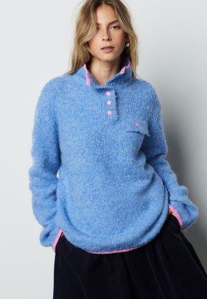 Frau trägt einen flauschigen blauen Pullover mit pinken Knöpfen und Besatz, steht mit den Händen in den Taschen vor einem schlichten hellen Hintergrund.