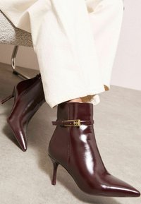 Burgundijas lakoti ādas potīšu zābaki ar smailu purngalu, dekoratīva siksna un zelta toņa sprādze, uz neitrāla fona.