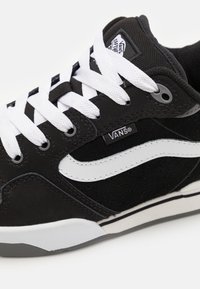 Sneaker din suede negru cu accente albe, având sireturi groase albe, o guler căptușit și o talpă din cauciuc texturat. Logo-ul este vizibil pe limba încălțămintei.