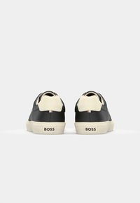 Adidași din piele negri și crem cu vârful rotunjit, accente contrastante la călcâi și branding "BOSS" pe spate. Textură netedă.