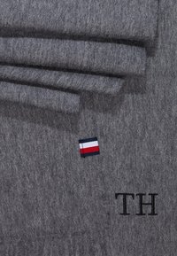 Szara filcowa tkanina o miękkiej fakturze, z małym wyhaftowanym logo w kolorach czerwonym, białym i niebieskim oraz inicjałami "TH" w czarnym ściegu.