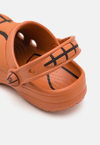 Sabot portocaliu din cauciuc cu accente negre, având o curea rotundă, orificii de ventilație și o talpă texturată cu un design sportiv.