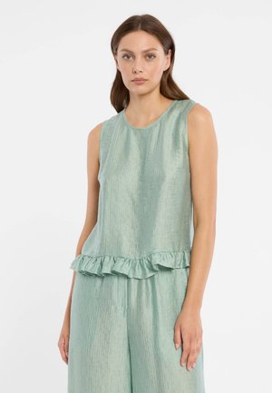 Vrouw met bruin haar die een mouwloze lichtgroene top met ruches en bijpassende wijde broek draagt, tegen een effen witte achtergrond.
