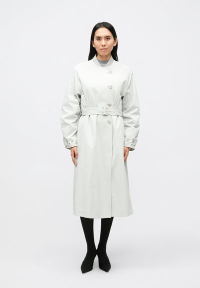 Hellgrauer Trenchcoat mit Gürtel, Knopfleiste und großen Manschetten. Verfügt über eine gerade Silhouette und reicht bis zur Mitte der Wade. Wird über schwarzen Strumpfhosen getragen.