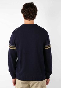 Pull en maille bleu marine avec un col rond et des manches longues, arborant trois rayures horizontales en or et gris sur le haut des bras.