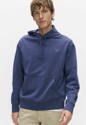 Jonge man die een blauwe capuchontrui met voorzak en beige broek draagt, staand tegen een effen lichte achtergrond.