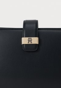 Melna ādas rokassoma ar zeltītu aizdari un iegravētu logo. Dizains ir strukturēts ar gludu tekstūru un akurātu šuvju izpildījumu.