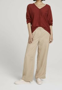 Pull en V rouge avec plis à l'avant, associé à un pantalon en lin beige ample et des baskets de couleur claire. Tissu texturé, coupe décontractée.