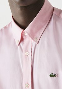 Camicia a collare rosa chiaro con patta dei bottoni, caratterizzata da bottoni bianchi e un piccolo logo di coccodrillo verde ricamato sul petto.