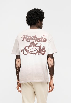 T-shirt à manches courtes de couleur crème clair avec un grand motif brun foncé indiquant « Radiate the Sole » au dos, comprenant des dessins abstraits et des accents.